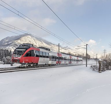 Ein roter Zug fährt im Winter durch verschneite Landschaften, vorbei an einer kleinen Kapelle, mit Bergen im Hintergrund unter einem klaren, blauen Himmel.