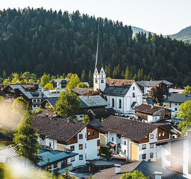 Panoramic view of the Wilder Kaiser mountain range with a picturesque village in the foreground, lush green forests, and clear blue skies.