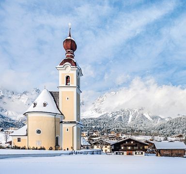 Snowy village with a church near the Wilder Kaiser mountains under a blue sky.