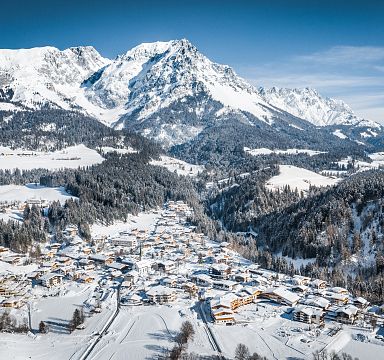 Verschneites Bergdorf in Tirol mit traditionellen Häusern, umgeben von beeindruckenden Alpenbergen unter blauem Himmel. Winterliche, idyllische Landschaft.