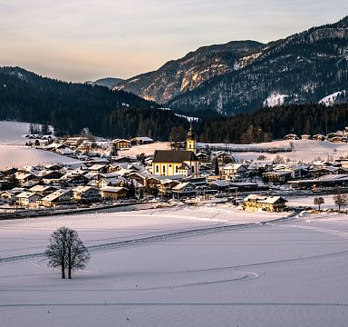 A snow-covered village nestled in the Wilder Kaiser region, with mountains in the background and warm sunlight illuminating the landscape.