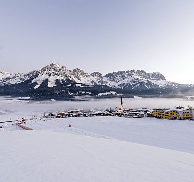 Winterliches Alpenpanorama mit verschneiten Bergen und einem ruhigen Tal, im Vordergrund ein Dorf mit Kirche und traditionellen Gebäuden unter blauem Himmel.