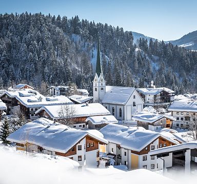 Winterliche Berglandschaft mit verschneiten Häusern und Kirche in einem Dorf, umgeben von bewaldeten Hügeln und Bergen unter klarem Himmel.