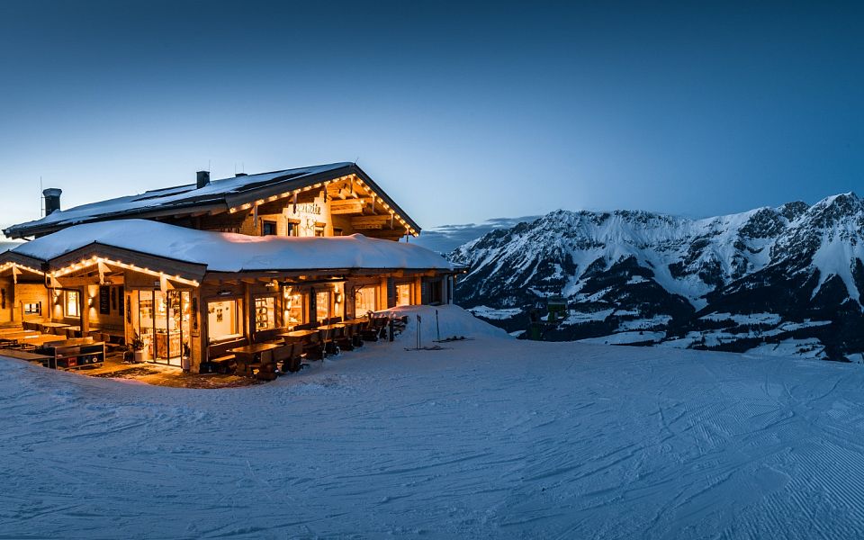 A serene winter scene at Wilder Kaiser with snow-covered trees and mountains framing a peaceful village under a warm sunrise.