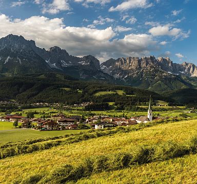 Panorama der Region Wilder Kaiser mit majestätischem Bergmassiv im Hintergrund, grünen Wiesen im Vordergrund und einem Dorf mit einer markanten Kirche.