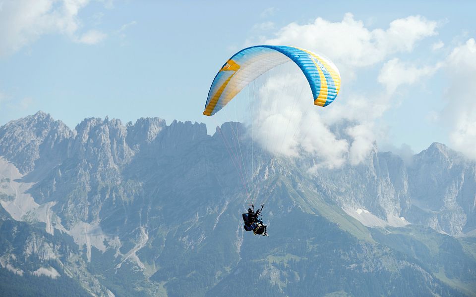 A paraglider is seen gliding over the Wilder Kaiser mountains, with clear skies and rugged peaks in the background, showcasing the beauty of Tyrol.