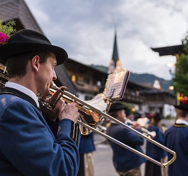 Blaskapelle in traditioneller Tracht spielt auf einer Straße, während Kirchturm im Hintergrund zu sehen ist. Die Musiker tragen Hüte mit Blumen.