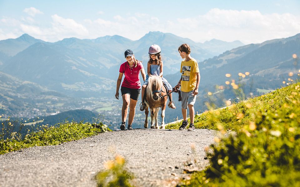 Drei Kinder begleiten ein Pony auf einem sonnigen Wanderweg in den Bergen. Im Hintergrund sind grüne Hänge und blaue Himmel mit ein paar Wolken zu sehen.