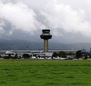 Airport control tower with planes parked adjacent, set against a cloudy mountain backdrop and green field in the foreground.