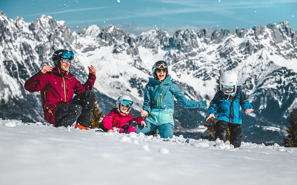 A family plays in the snow on a sunny day in the Wilder Kaiser region, with snow-capped mountains in the background and clear blue skies.