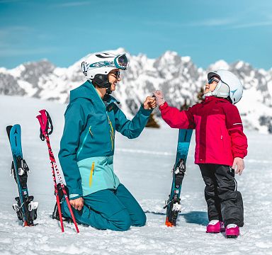 Erwachsener und Kind im Schnee mit Skiausrüstung vor beeindruckendem Alpenpanorama, lächeln sich an. Die Szene wirkt winterlich und fröhlich.