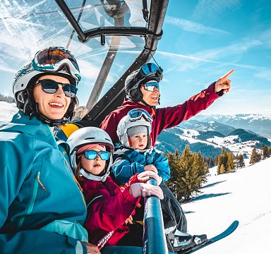 Familie in Skikleidung sitzt auf einem Sessellift in schneebedeckter Berglandschaft. Kinder tragen Helme, während der Erwachsene in die Ferne zeigt.