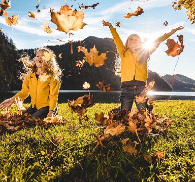 Two children playfully throwing autumn leaves in the air on a grassy field with mountains and a clear sky in the background.