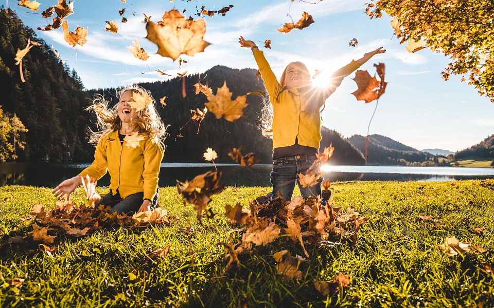 Two children playfully throwing autumn leaves in the air on a grassy field with mountains and a clear sky in the background.