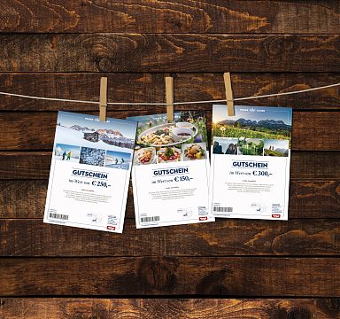 Drei Gutscheine hängen mit Klammern an einer Leine vor einer rustikalen Holzwand. Jeder Gutschein zeigt verschiedene Fotos und Werte in Euro.