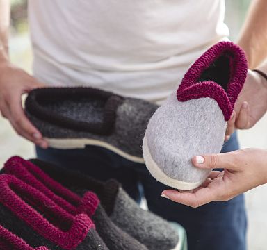 Personen halten warme, handgefertigte Filzhausschuhe in Grau und Rot. Ideal für kalte Tage. Detailaufnahme von Händen und Schuhen auf einem Markt.