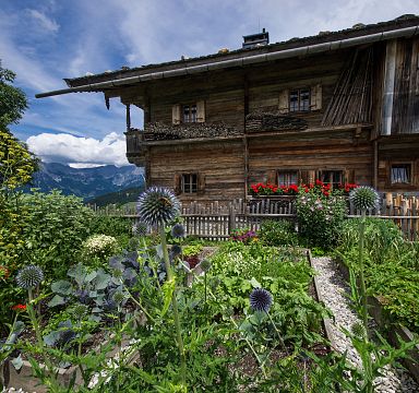 Ein traditionelles alpines Holzhaus mit üppigem Garten voller bunter Blumen und Gemüse, umgeben von Berglandschaft unter blauem Himmel.