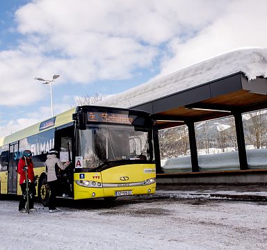 A bus parked at a snow-covered station in the Wilder Kaiser region, with people in winter gear preparing to board. Snow-capped mountains can be seen in the background.