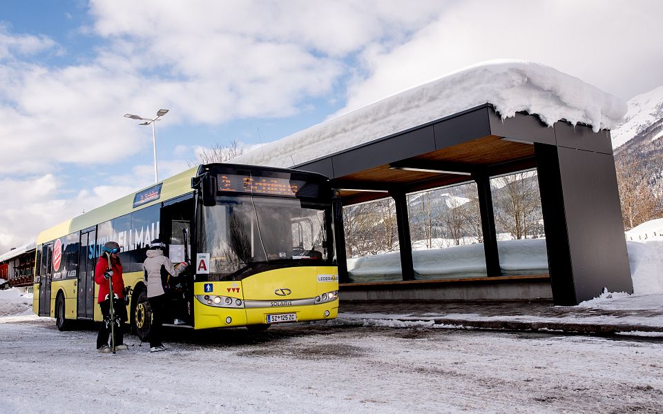 Ein gelber Bus hält an einer verschneiten Haltestelle. Eine Person steigt mit Skiern ein. Im Hintergrund verschneite Berge unter einem bewölkten Himmel.