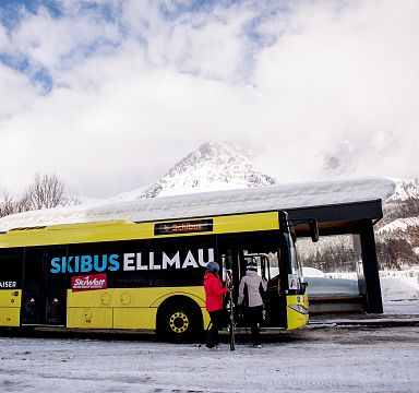 Ein gelber Skibus mit der Aufschrift "SKIBUS ELLMAU" steht an einer Bushaltestelle im Schnee. Eine Person steigt ein, umgeben von schneebedeckter Landschaft.