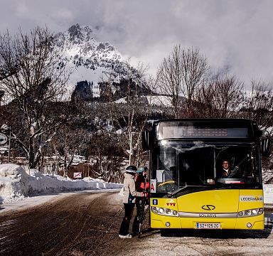 Ein gelber Bus hält an einer verschneiten Haltestelle in einer Bergregion. Ein Skifahrer steigt ein, im Hintergrund sind schneebedeckte Berge zu sehen.