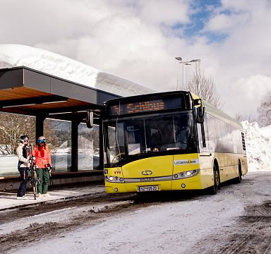 Ein gelber Bus steht an einer Haltestelle in einer verschneiten Landschaft. Zwei Personen mit Wintersportausrüstung gehen zum Bus. Der Himmel ist bewölkt.