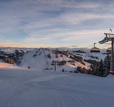 Verschneite Berghänge mit Skiliften unter blauem Himmel, umgeben von Tannenbäumen, bieten einen idyllischen Anblick einer Wintersportregion in den Alpen.