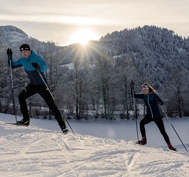 Zwei Menschen beim Langlauf in einer verschneiten Berglandschaft. Die Sonne geht hinter den schneebedeckten Hügeln auf und der Himmel ist leicht bewölkt.