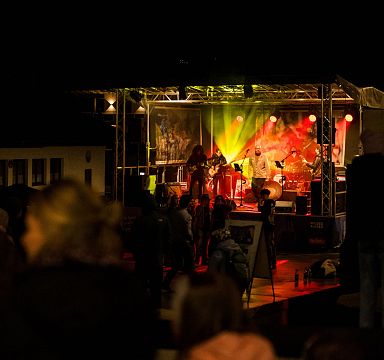 Eine nächtliche Konzertbühne im Freien, beleuchtet von bunten Lichtern. Menschen stehen davor und lauschen der Musik in einer entspannten Atmosphäre.