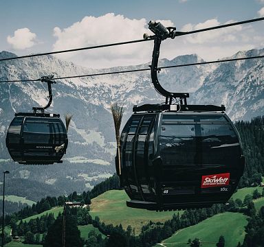 Cable cars traverse a verdant landscape, with towering peaks of the Wilder Kaiser in the distance, under a partly cloudy sky.