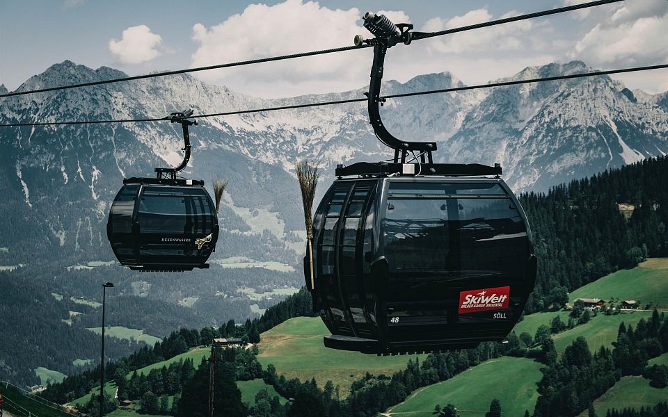 Zwei moderne Gondeln schweben über grüne Hänge vor einer beeindruckenden Bergkulisse in den Alpen, mit klarem Himmel und vereinzelten Wolken.