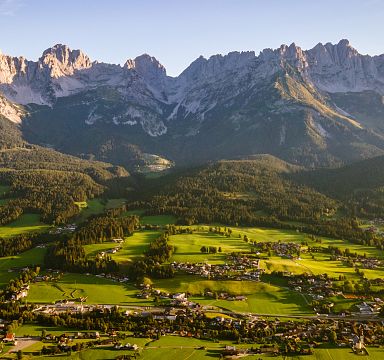 Panoramablick auf das imposante Bergmassiv des Wilden Kaisers in Tirol, eingerahmt von grünen Wiesen und malerischen Dörfern unter einem klaren blauen Himmel.