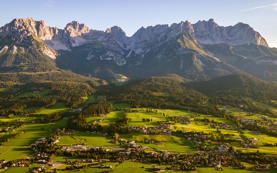 Panoramablick auf das imposante Bergmassiv des Wilden Kaisers in Tirol, eingerahmt von grünen Wiesen und malerischen Dörfern unter einem klaren blauen Himmel.