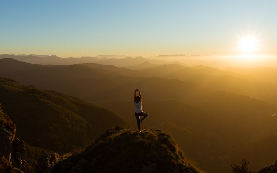 Person steht in Yoga-Position auf einem Berggipfel bei Sonnenuntergang, umgeben von hügeliger Landschaft und goldenem Licht des Abendhimmels.