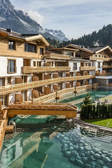View of a luxury resort in the Wilder Kaiser, featuring traditional wooden architecture, a crystal-clear alpine pool, and mountainous backdrop on a sunny day.