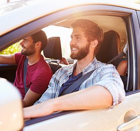 Ein Auto fährt eine Landstraße entlang, umgeben von grünen Wiesen und Bäumen. Im Hintergrund sind schneebedeckte Berge unter einem blauem Himmel zu sehen.