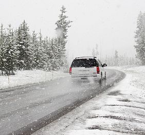 Ein Auto fährt auf einer schneebedeckten Landstraße durch einen verschneiten Wald, während es stark schneit. Die Szenerie wirkt kalt und winterlich.