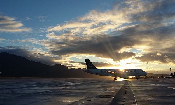 Ein Flugzeug rollt bei Sonnenuntergang auf dem Rollfeld. Die Sonne strahlt durch dichte Wolken und erzeugt eine stimmungsvolle Atmosphäre mit Bergsilhouette.