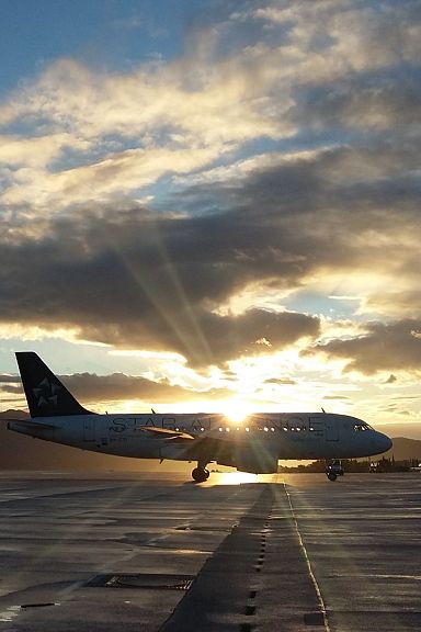 Ein Flugzeug rollt bei Sonnenuntergang auf dem Rollfeld. Die Sonne strahlt durch dichte Wolken und erzeugt eine stimmungsvolle Atmosphäre mit Bergsilhouette.