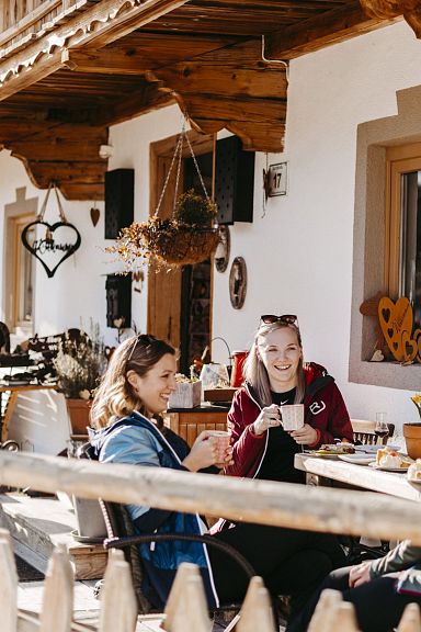 Three people sit at a table outside a rustic hut with skis nearby, enjoying food and sunshine. The scene is set in the picturesque Wilder Kaiser area.
