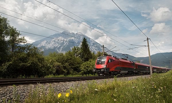 Ein roter Zug fährt auf Gleisen durch eine grüne Landschaft, im Hintergrund erhebt sich ein bewaldetes Bergmassiv unter einem bewölkten Himmel.