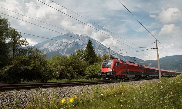 Eine rote, moderne Lok fährt durch grüne Felder vor einem Gebirgszug, unter einem blauen Himmel mit weißen Wolken.