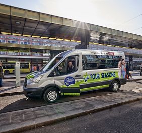 Ein grüner Shuttlebus mit der Aufschrift "Four Seasons" steht auf einer städtischen Straße, im Hintergrund sind moderne Gebäude und eine Bushaltestelle zu sehen.