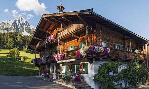 Wooden chalet with colorful flowers and Wilder Kaiser mountains in the background, under a clear blue sky.