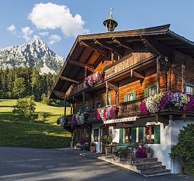 Ein traditionelles Tiroler Holzhaus mit üppigen Blumenbalkonen vor einem grünen Hügel und einem majestätischen Bergmassiv im Hintergrund.
