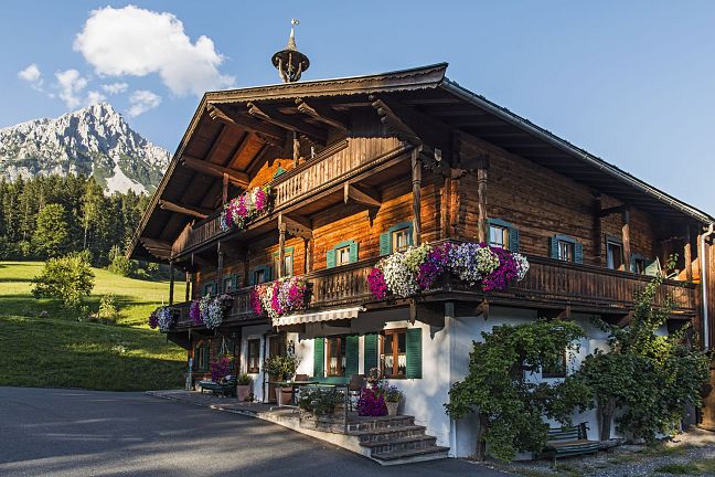 Ein traditionelles Tiroler Holzhaus mit üppigen Blumenbalkonen vor einem grünen Hügel und einem majestätischen Bergmassiv im Hintergrund.