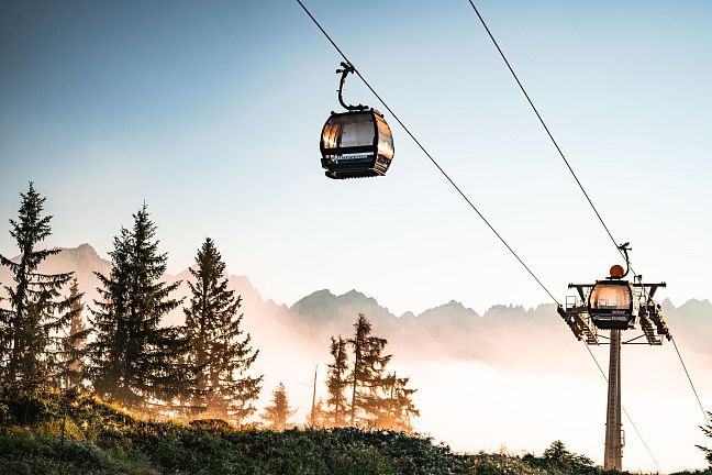 Cable cars ascending with the majestic Wilder Kaiser mountains in the background, surrounded by trees and a hazy sky at sunrise.
