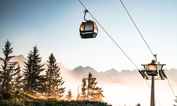 Cable cars ascending with the majestic Wilder Kaiser mountains in the background, surrounded by trees and a hazy sky at sunrise.