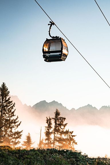 Cable cars ascending with the majestic Wilder Kaiser mountains in the background, surrounded by trees and a hazy sky at sunrise.