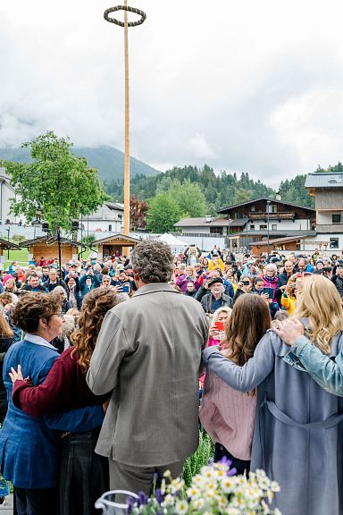 Eine große Menschenmenge auf einem Dorfplatz, umgeben von Bergen. Im Vordergrund umarmen sich mehrere Personen und schauen in die Menge.